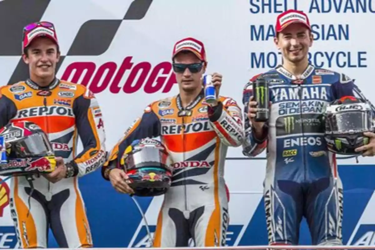 Tres pilotos de motociclismo en el podio sosteniendo cascos y latas de bebida energética, con trajes de carreras de sus respectivos equipos.