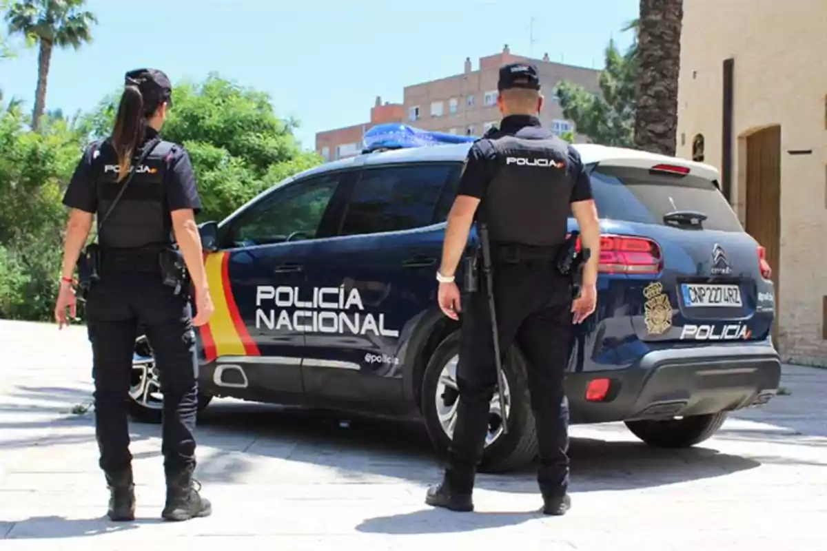 Dos agentes de la Policía Nacional de pie junto a un coche patrulla en una zona urbana.