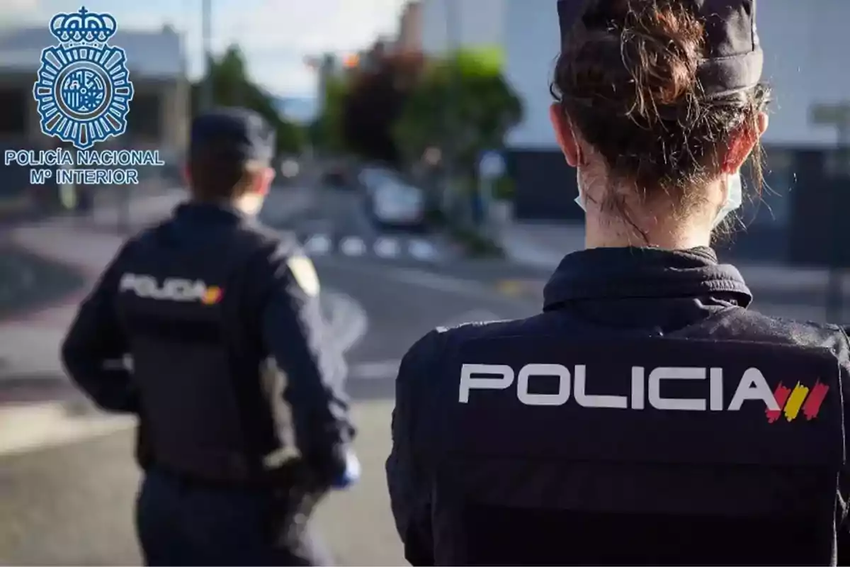 Dos agentes de la Policía Nacional de España de espaldas en una calle, con el logotipo de la Policía Nacional en la esquina superior izquierda.