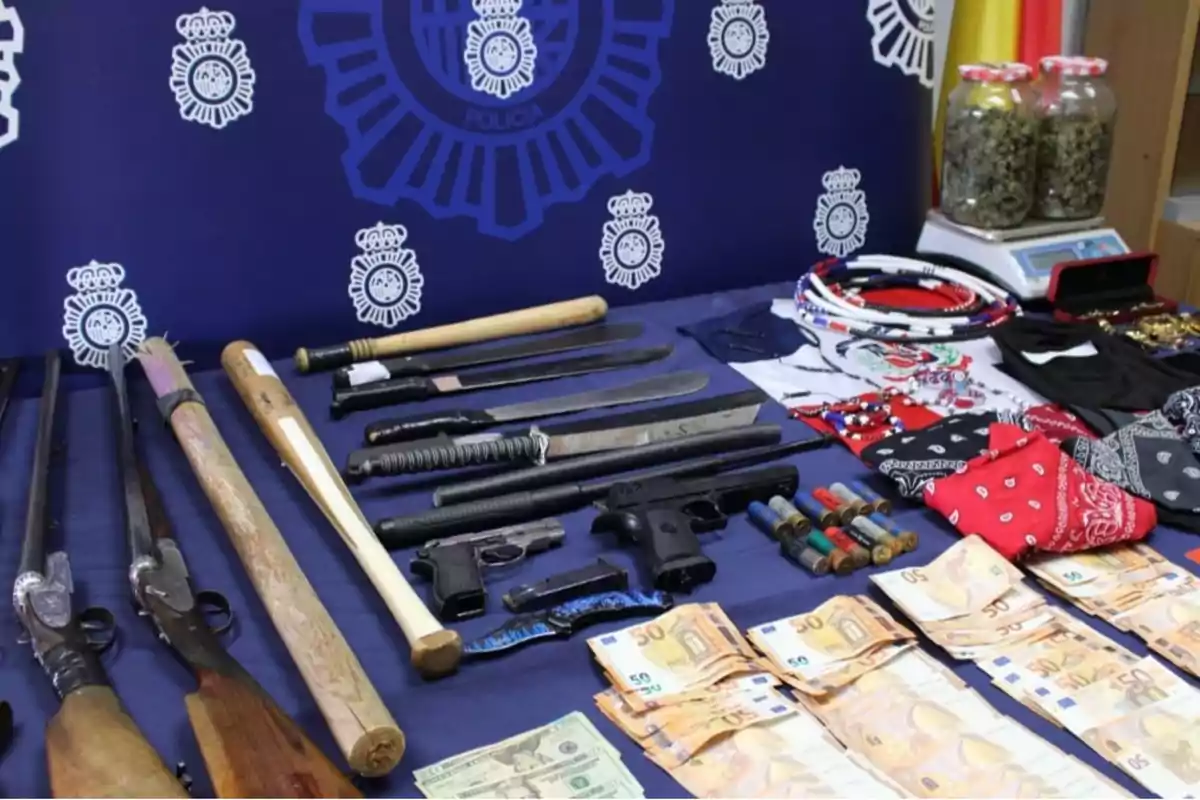 Mesa con armas, dinero, drogas y otros objetos incautados frente a un fondo con el logo de la policía.