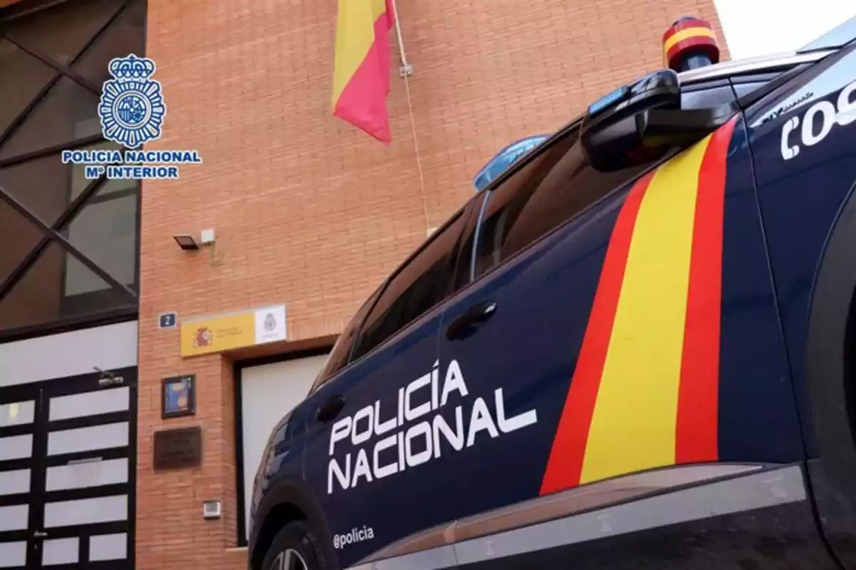Coche de la Policía Nacional de España estacionado frente a una comisaría con la bandera española ondeando y el logotipo oficial en la pared
