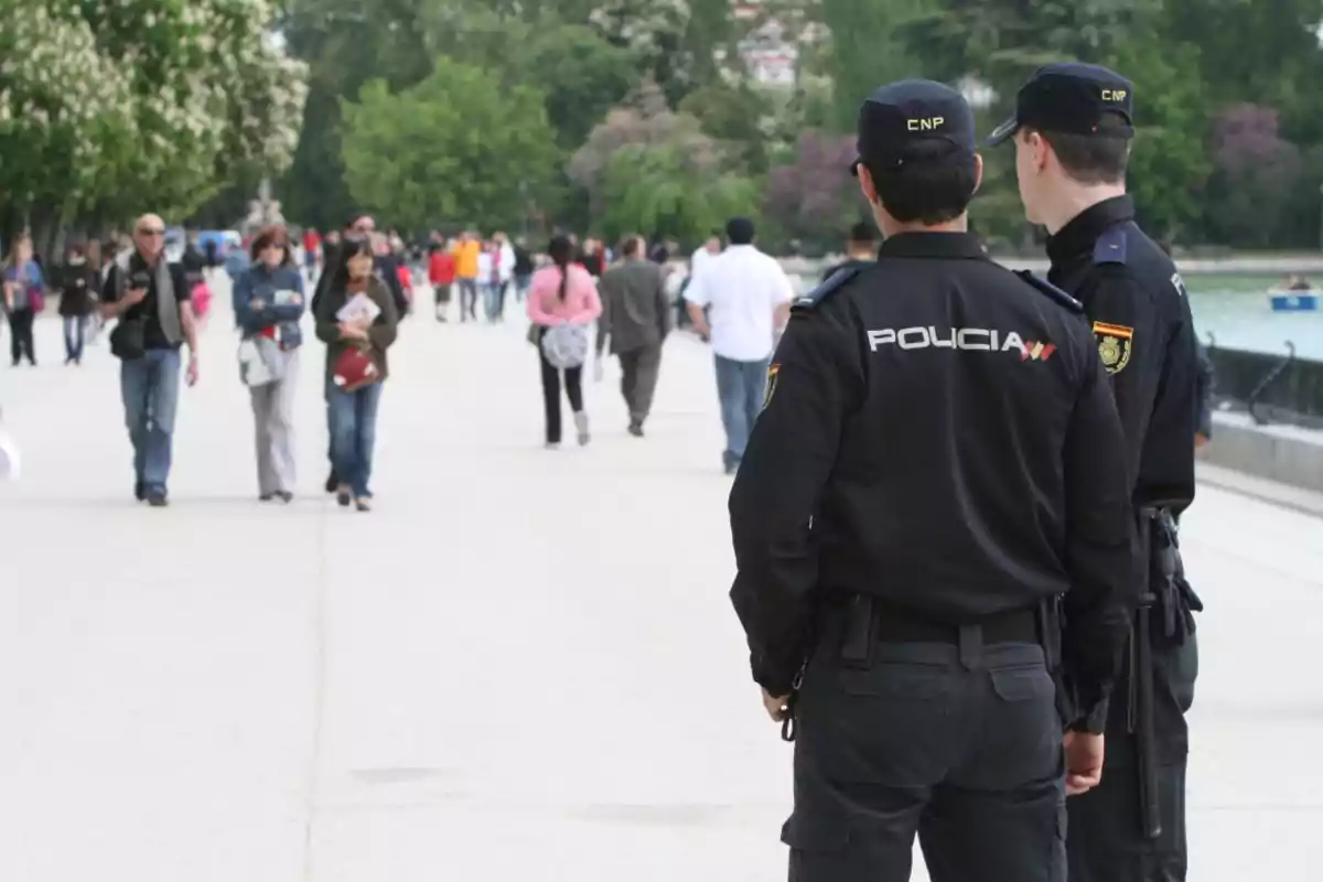 Dos policías de espaldas observan a un grupo de personas caminando por un paseo peatonal en un parque.