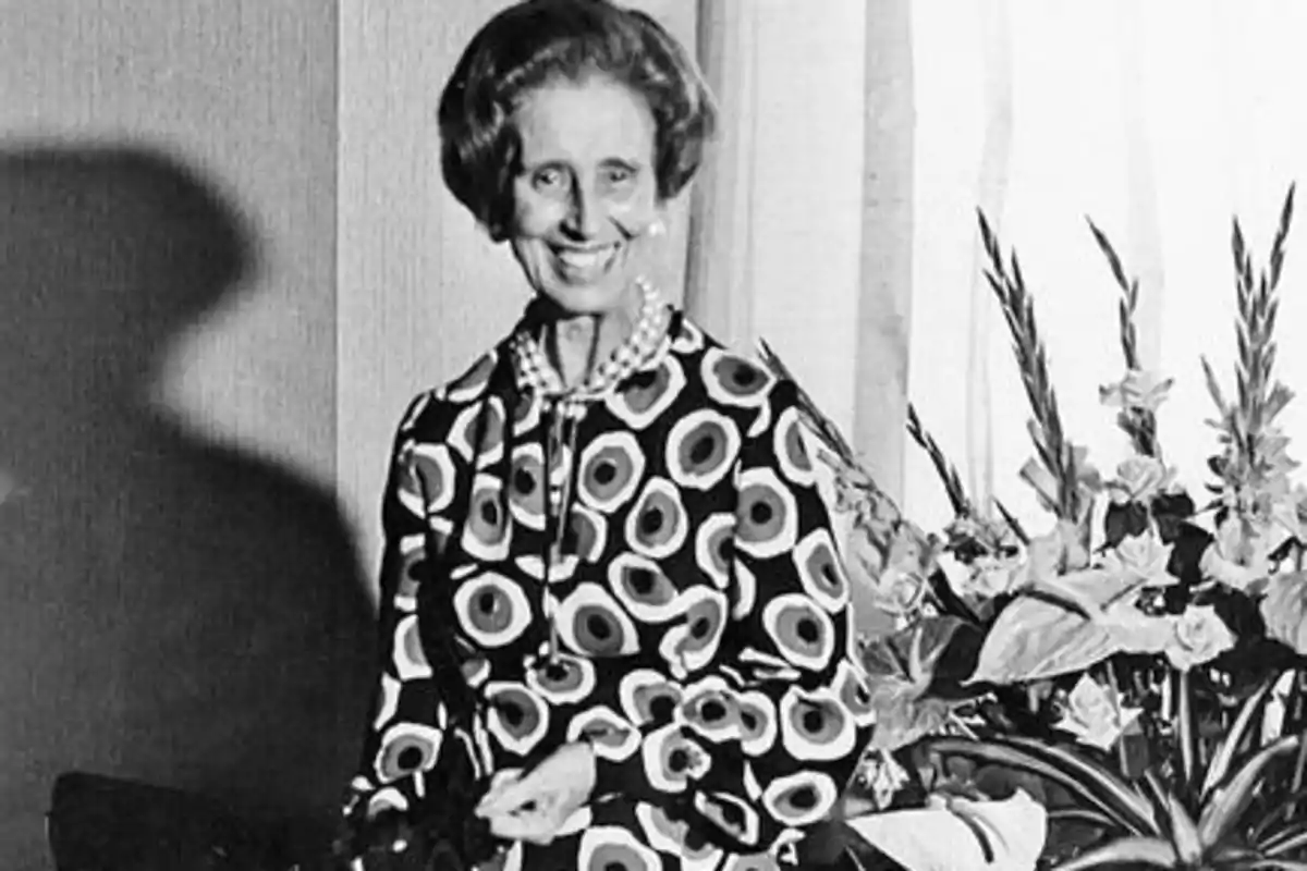 Mujer mayor sonriendo, vestida con un vestido de patrones circulares, con un collar de perlas y flores en el fondo.