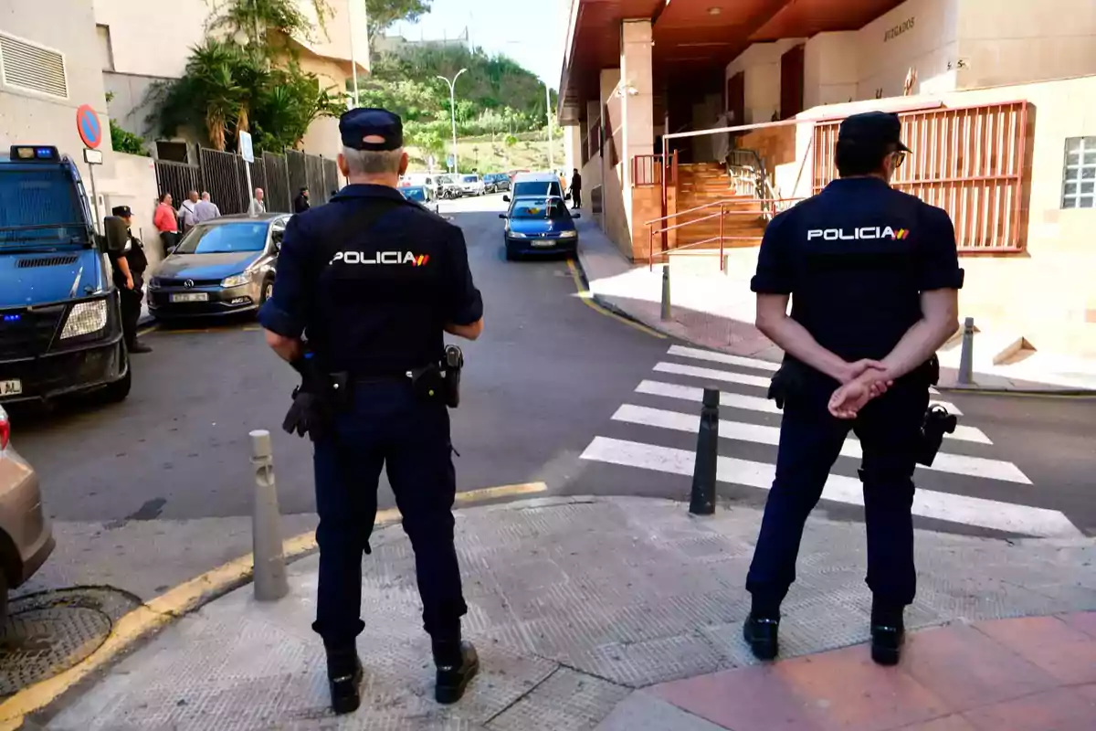 Dos policías de espaldas vigilando una calle cerca de un edificio con la palabra Dos policías de espaldas vigilando una calle cerca de un edificio con la palabra