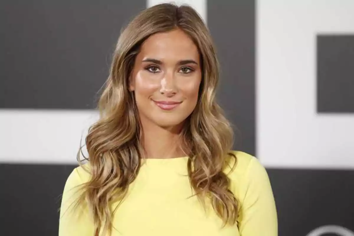 Mujer con cabello rubio ondulado y vestido amarillo sonriendo frente a un fondo con patrones geométricos.