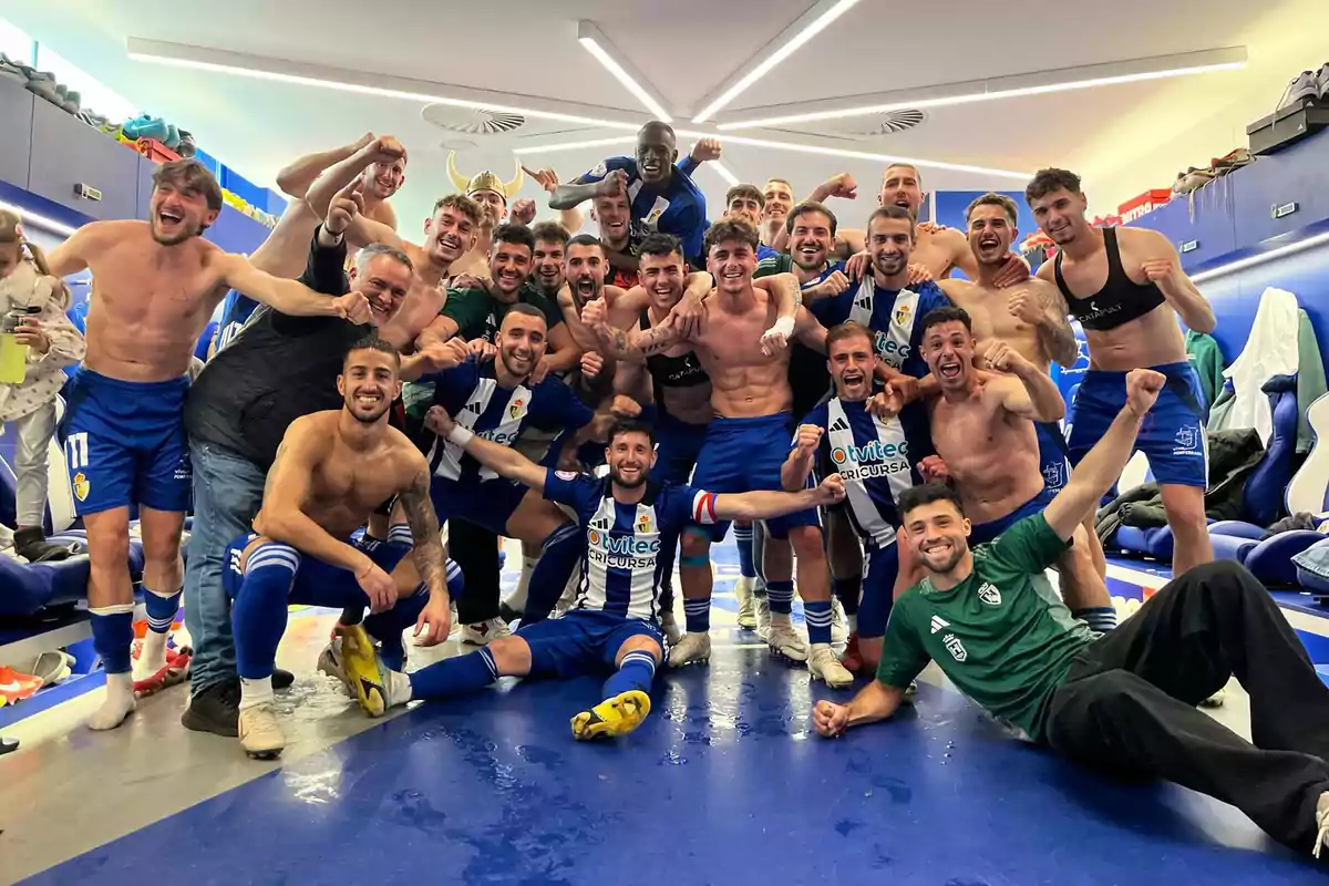Un grupo de jugadores de fútbol celebra con entusiasmo en el vestuario después de un partido, algunos están sin camiseta y todos muestran gestos de alegría y victoria.