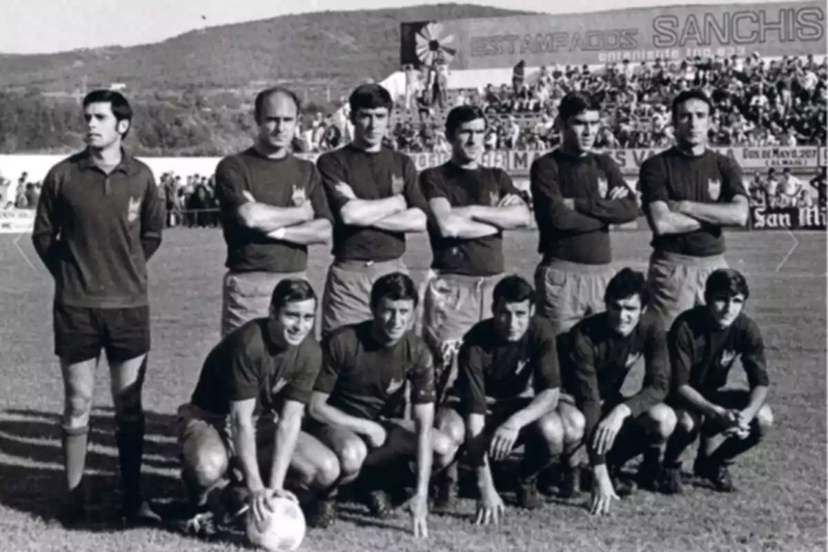 Un equipo de fútbol posando en el campo con una multitud en las gradas al fondo.