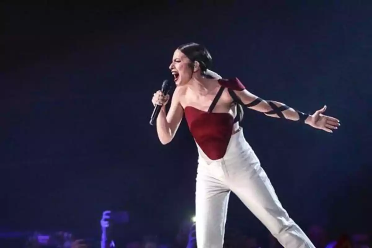 Cantante en el escenario con un micrófono, vestida con un top rojo y pantalones blancos, mientras canta apasionadamente.