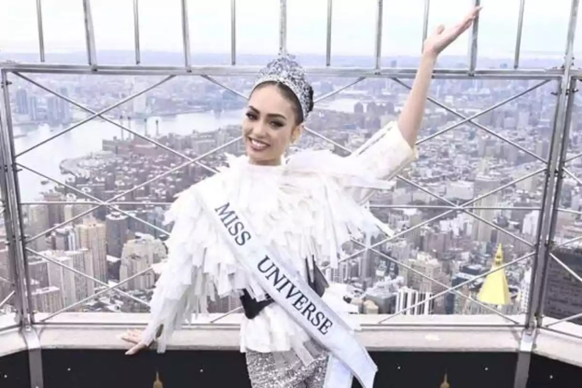 Persona con banda de "Miss Universe" y corona posando frente a una vista panorámica de la ciudad.