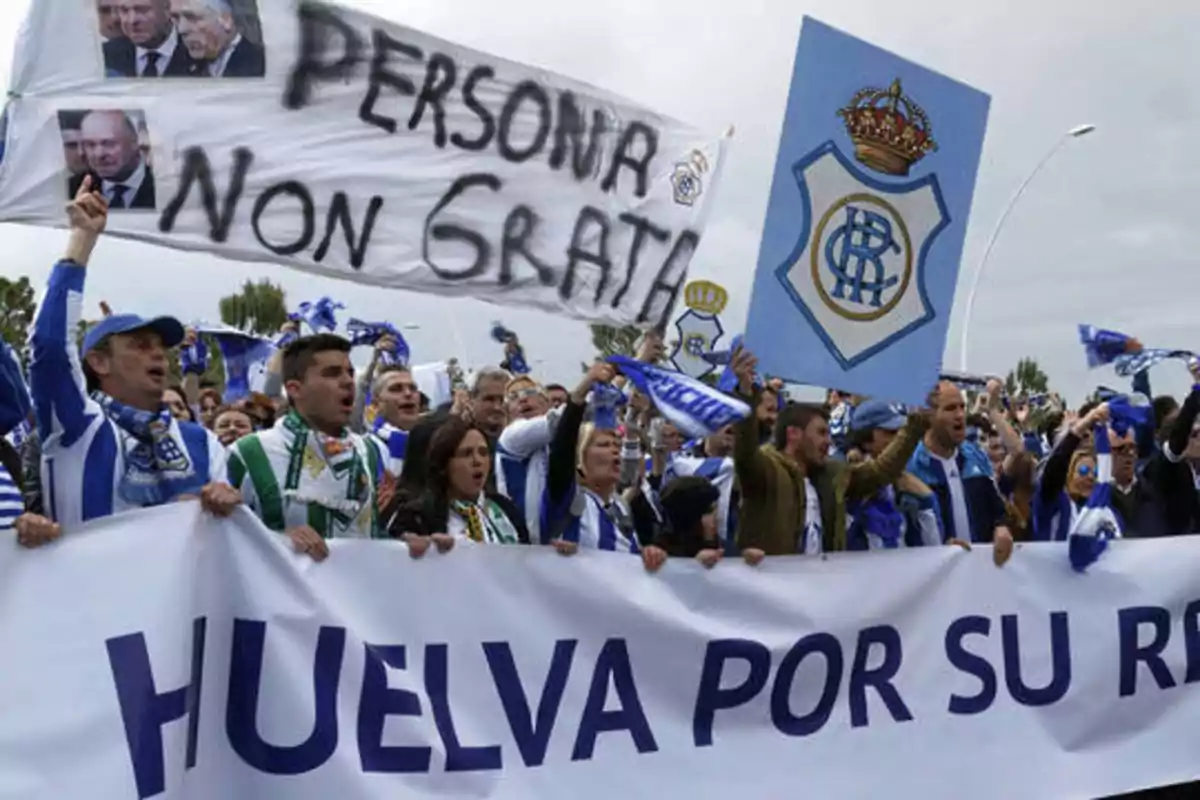 Un grupo de personas con bufandas y pancartas protesta en la calle, destacando un cartel que dice "Persona Non Grata" y otro con el escudo de un equipo de fútbol, mientras sostienen una gran pancarta que dice "Huelva por su RC".