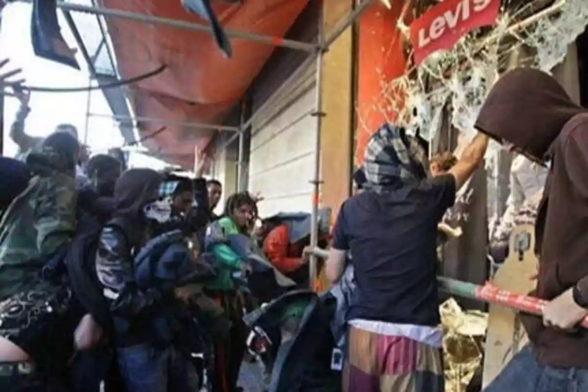 Un grupo de personas con capuchas y pañuelos en la cara rompiendo el escaparate de una tienda Levi's.