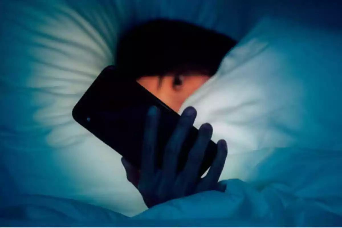 Persona acostada en la cama usando un teléfono móvil bajo las sábanas.