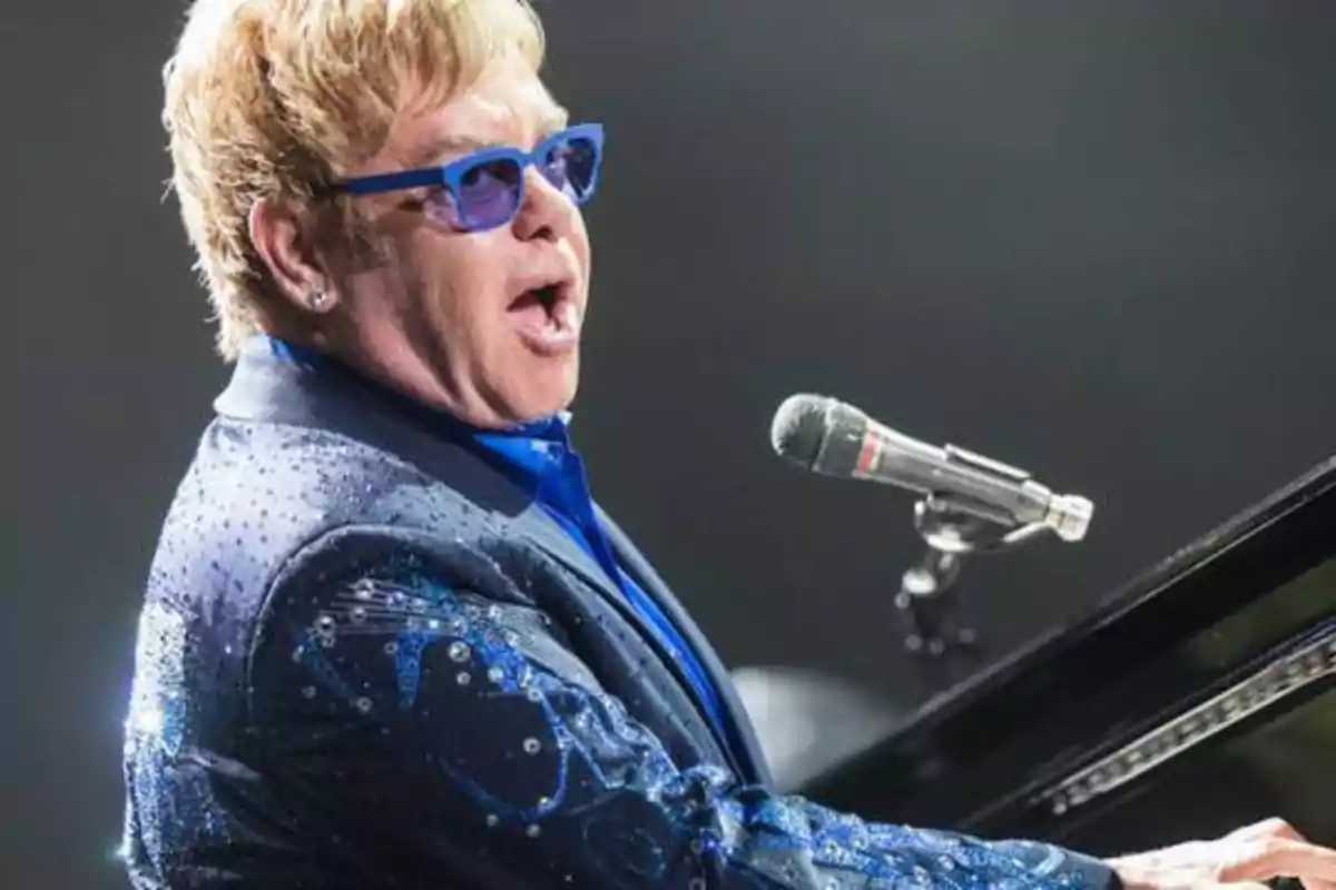 Una persona con gafas de sol azules y una chaqueta brillante está cantando y tocando el piano frente a un micrófono.