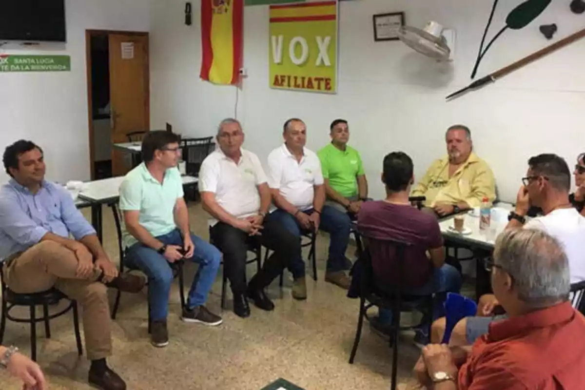 Un grupo de personas sentadas en una sala con una bandera de España y un cartel de VOX en la pared.