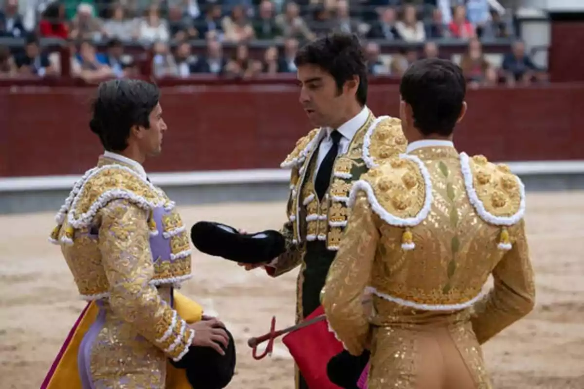 Tres toreros vestidos con trajes tradicionales de luces conversan en una plaza de toros, mientras el público observa desde las gradas.