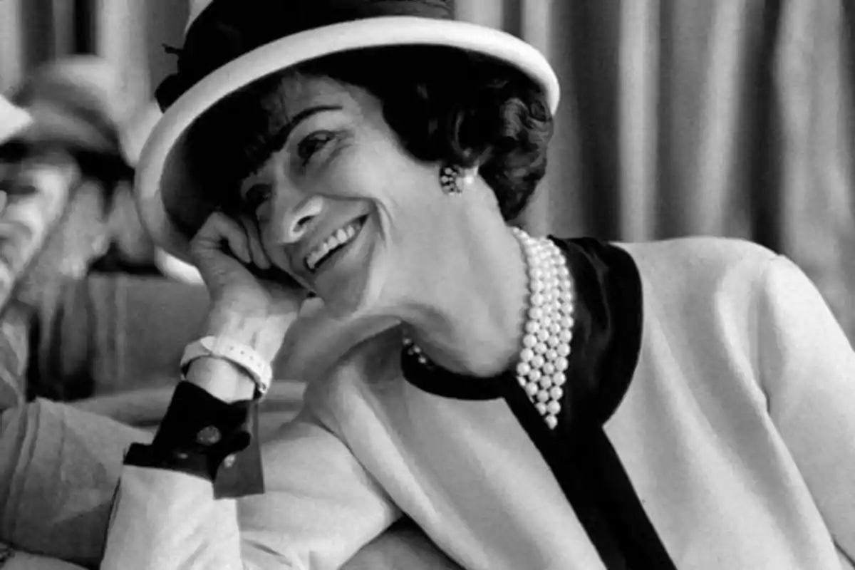 Mujer sonriendo con sombrero y collar de perlas.