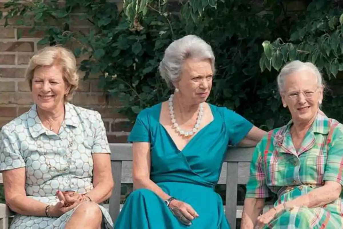 Tres mujeres mayores sentadas en un banco al aire libre, con una pared de ladrillo y vegetación de fondo.