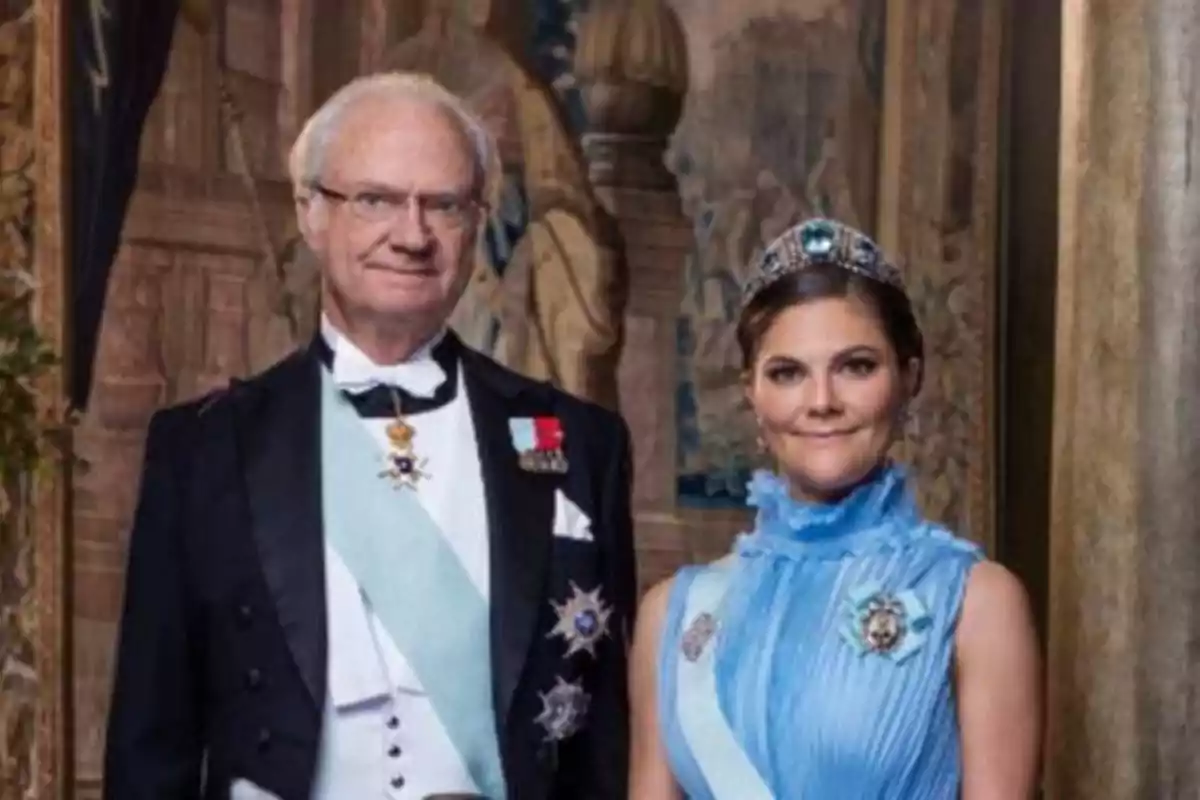 Una pareja vestida de gala, el hombre lleva un traje negro con condecoraciones y la mujer un vestido azul con una tiara.
