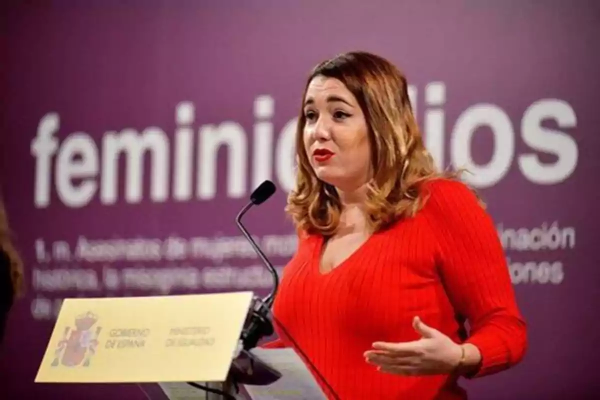 Mujer hablando en un podio con un cartel del Gobierno de España y el Ministerio de Igualdad, con la palabra "feminicidios" en el fondo.