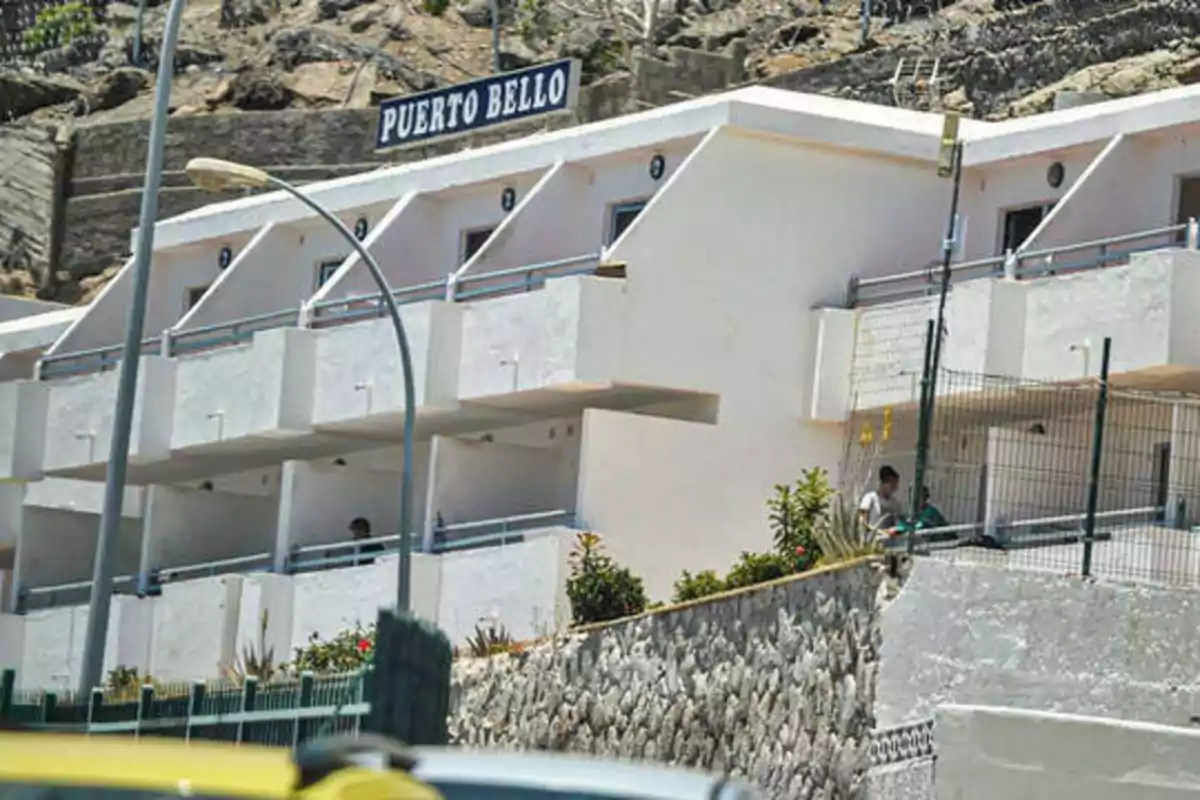 Edificio blanco de varios pisos con balcones y un letrero que dice "Puerto Bello" en la parte superior, ubicado en una zona montañosa con vegetación y una calle con una farola en primer plano.