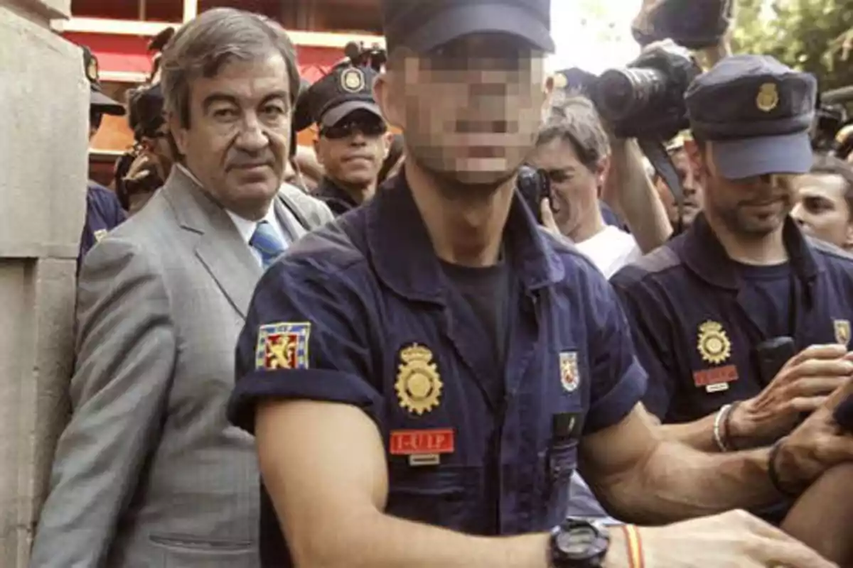 Un hombre de traje gris es escoltado por policías con uniformes azules mientras una multitud de fotógrafos y periodistas los rodea.