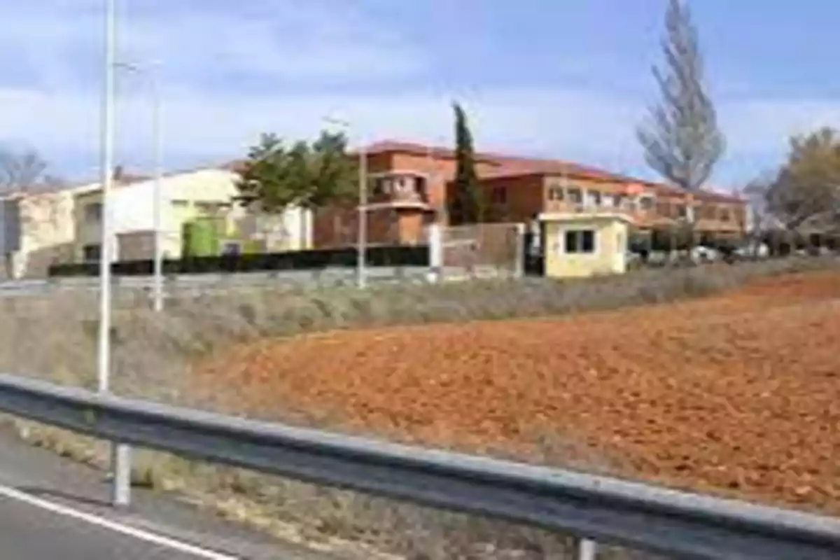 Edificios de ladrillo y cemento junto a una carretera con barandilla metálica y un campo de tierra en primer plano.