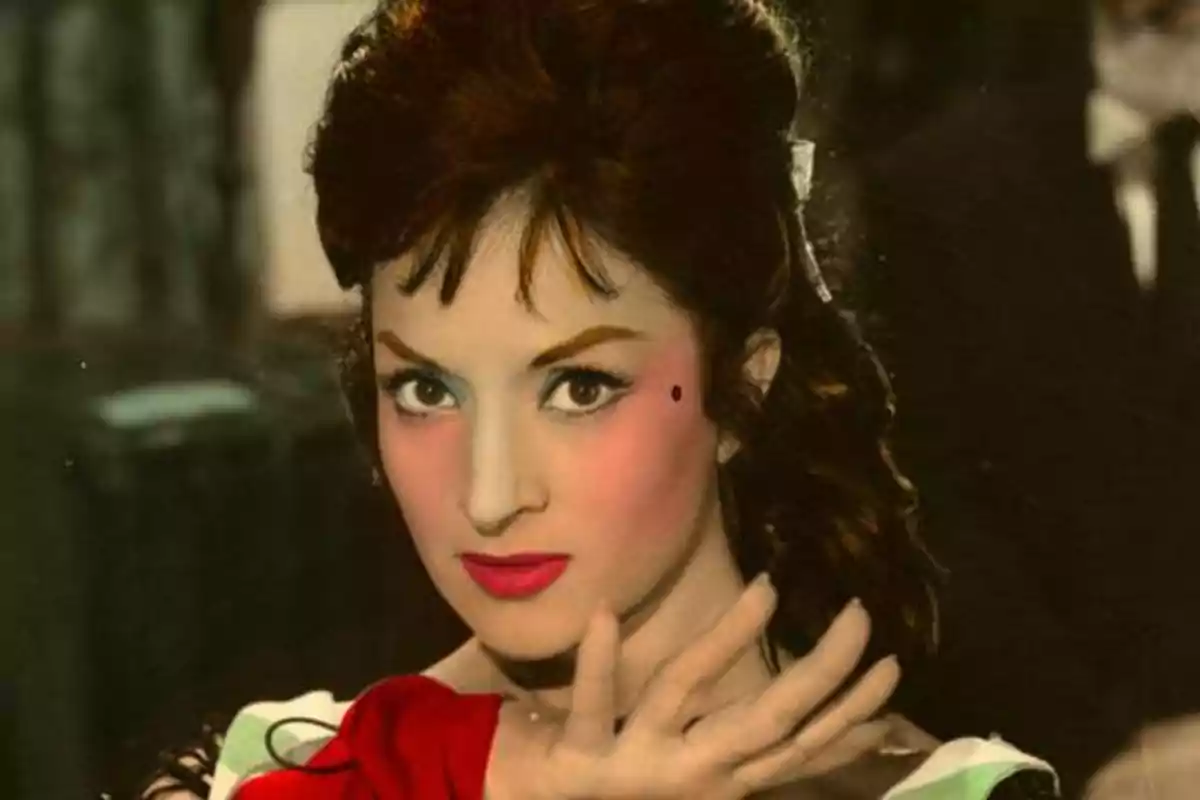 Una mujer con maquillaje llamativo y expresión seria, con la mano levantada frente a su rostro.