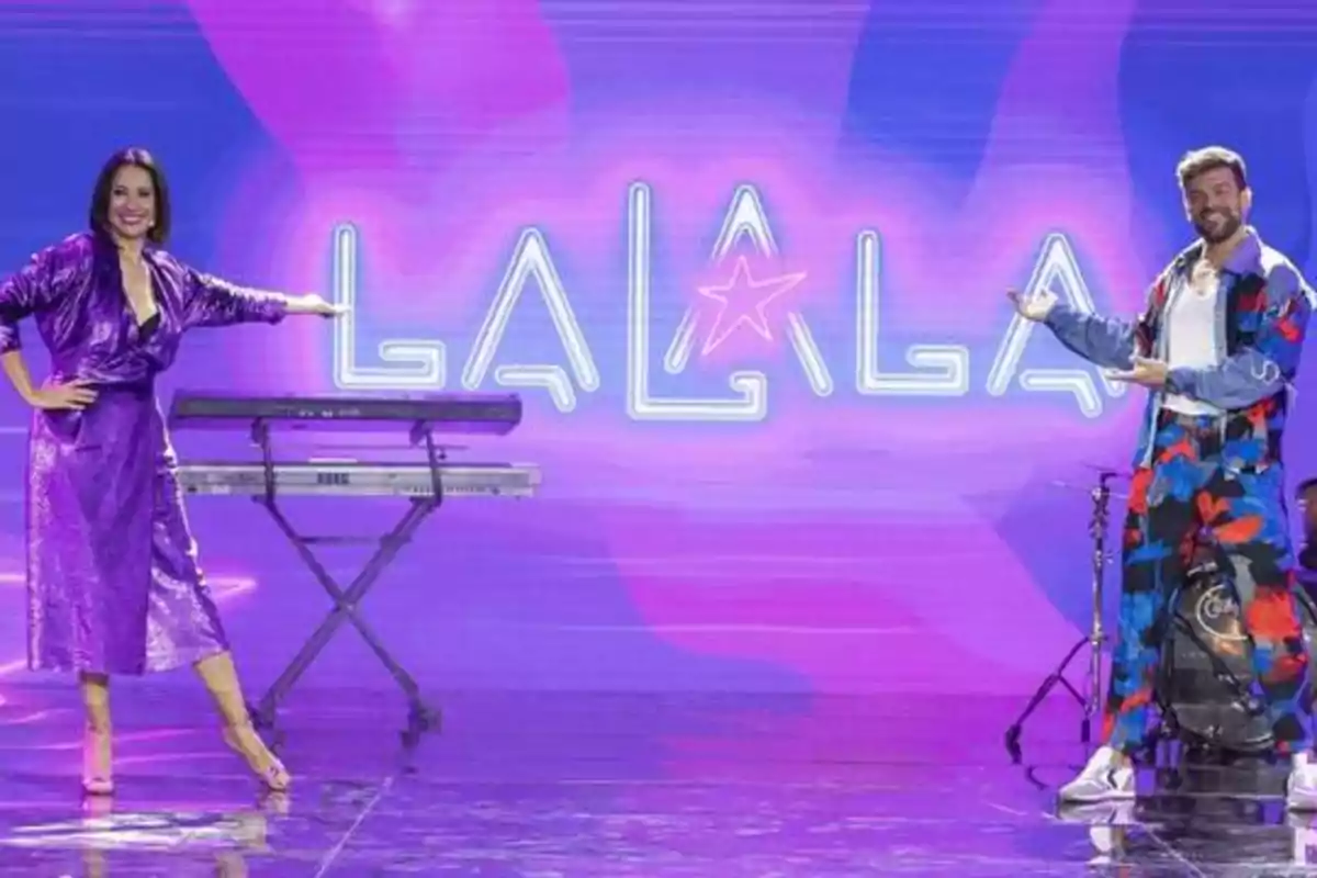Dos personas posan frente a un fondo con el texto "LALALA" iluminado en luces de neón, una de ellas está junto a un teclado y la otra junto a una batería.