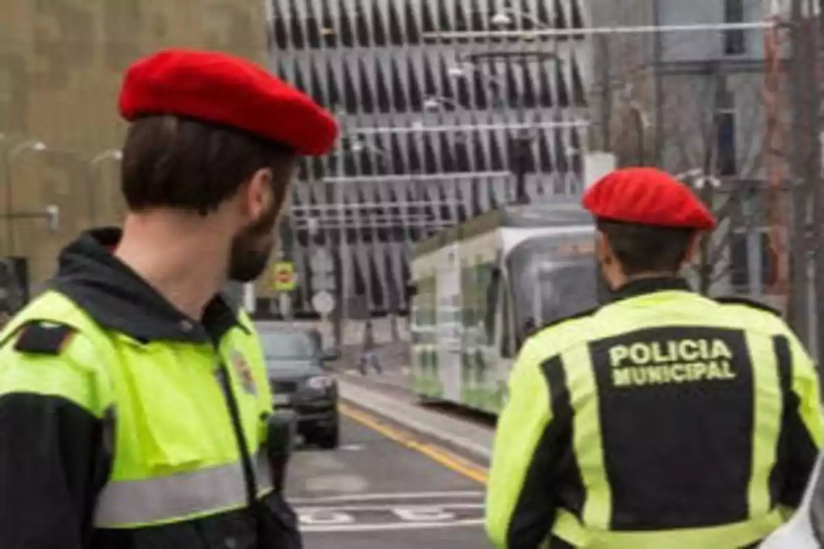 Dos policías municipales con boinas rojas y uniformes de alta visibilidad observan un tranvía en una calle urbana.