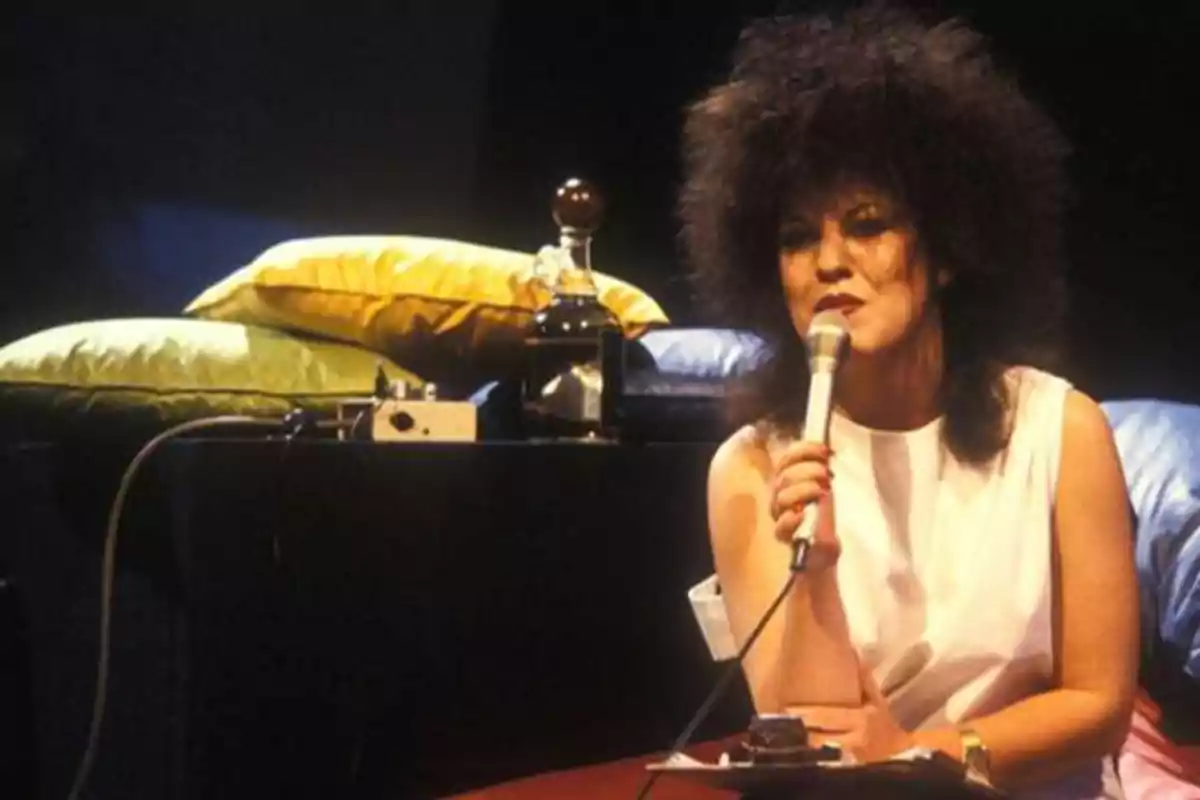 Persona con cabello rizado y voluminoso sosteniendo un micrófono, sentada frente a una mesa con cojines y equipo de audio.