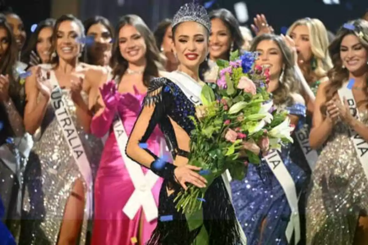Una mujer con una corona y un ramo de flores es felicitada por otras concursantes en un certamen de belleza.