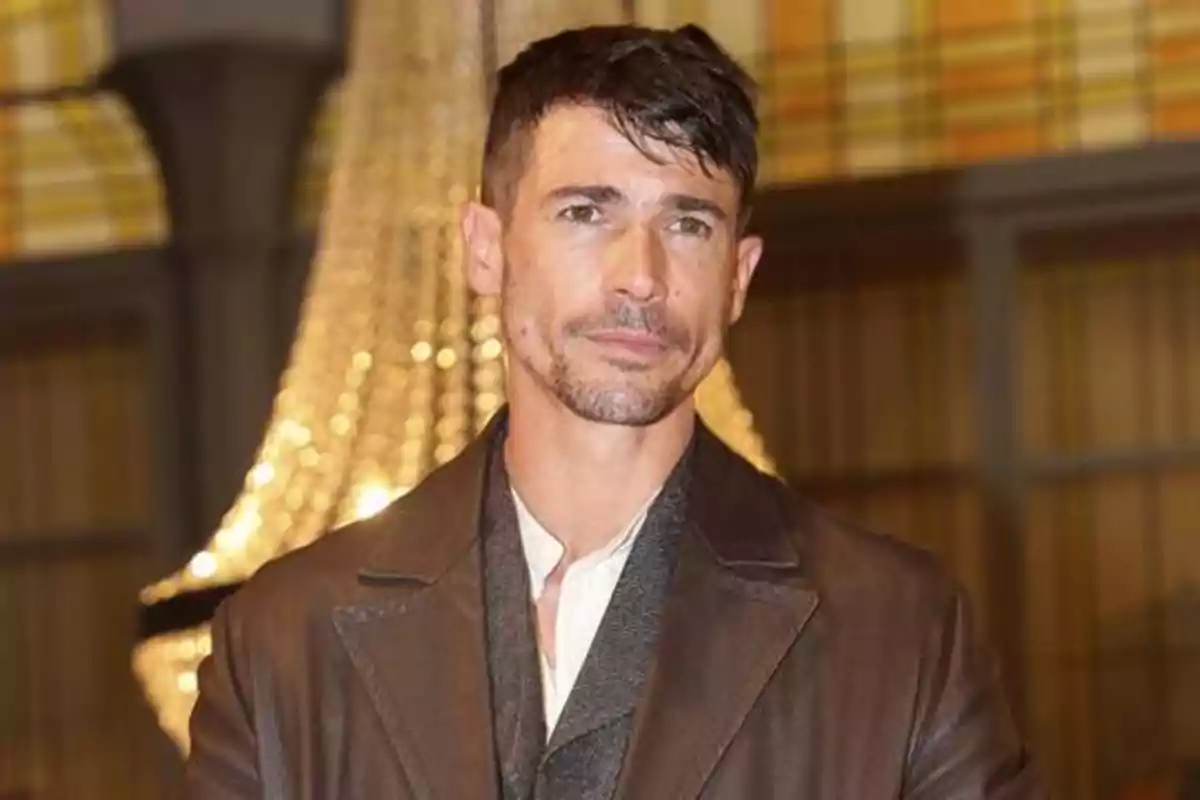 Hombre con chaqueta marrón posando frente a un fondo con luces brillantes.
