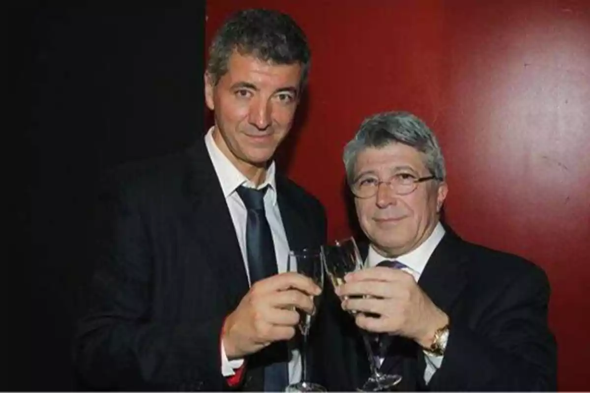 Dos hombres en trajes formales brindando con copas de champán frente a un fondo rojo.