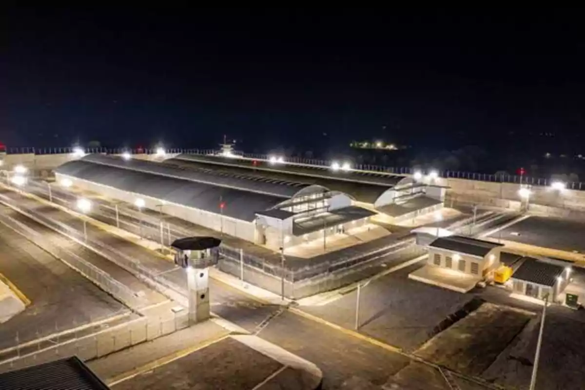 Vista aérea nocturna de una prisión iluminada con varias torres de vigilancia y edificios dentro de un perímetro cercado.
