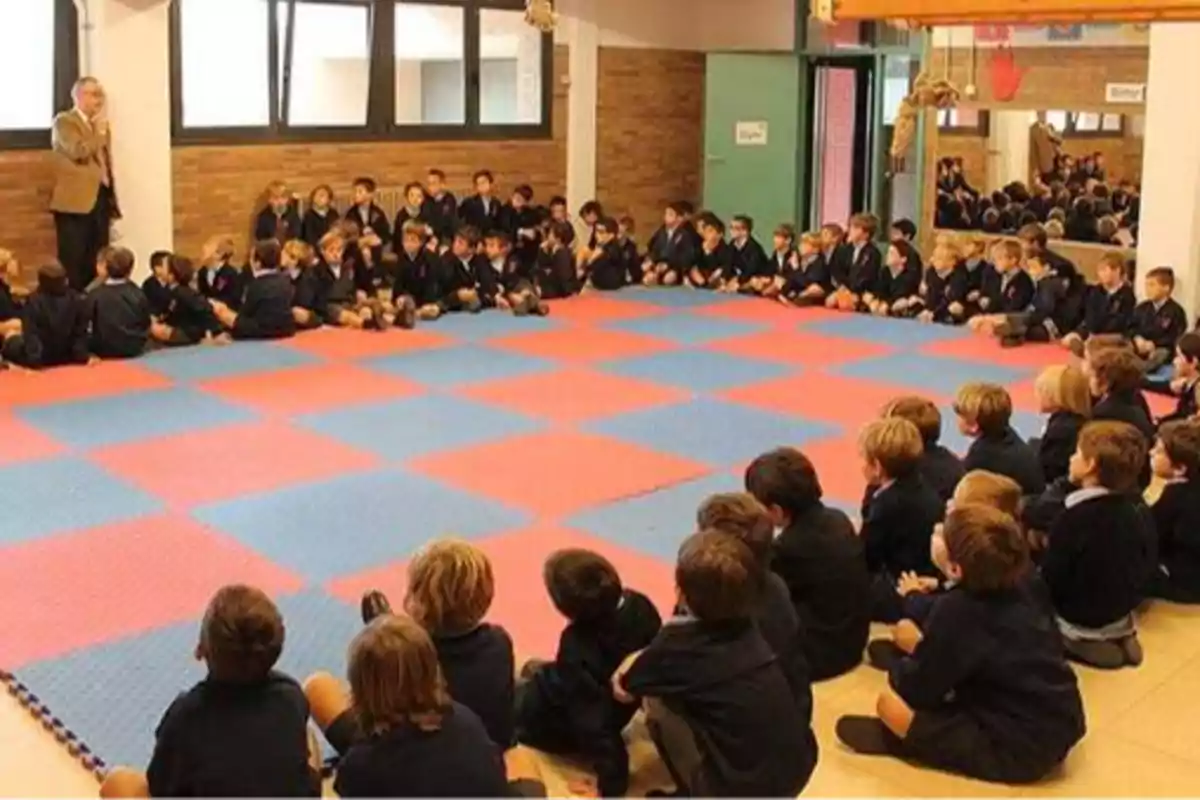 Un grupo de niños sentados en el suelo de una sala con alfombra de colores, escuchando a un hombre que está de pie hablando.