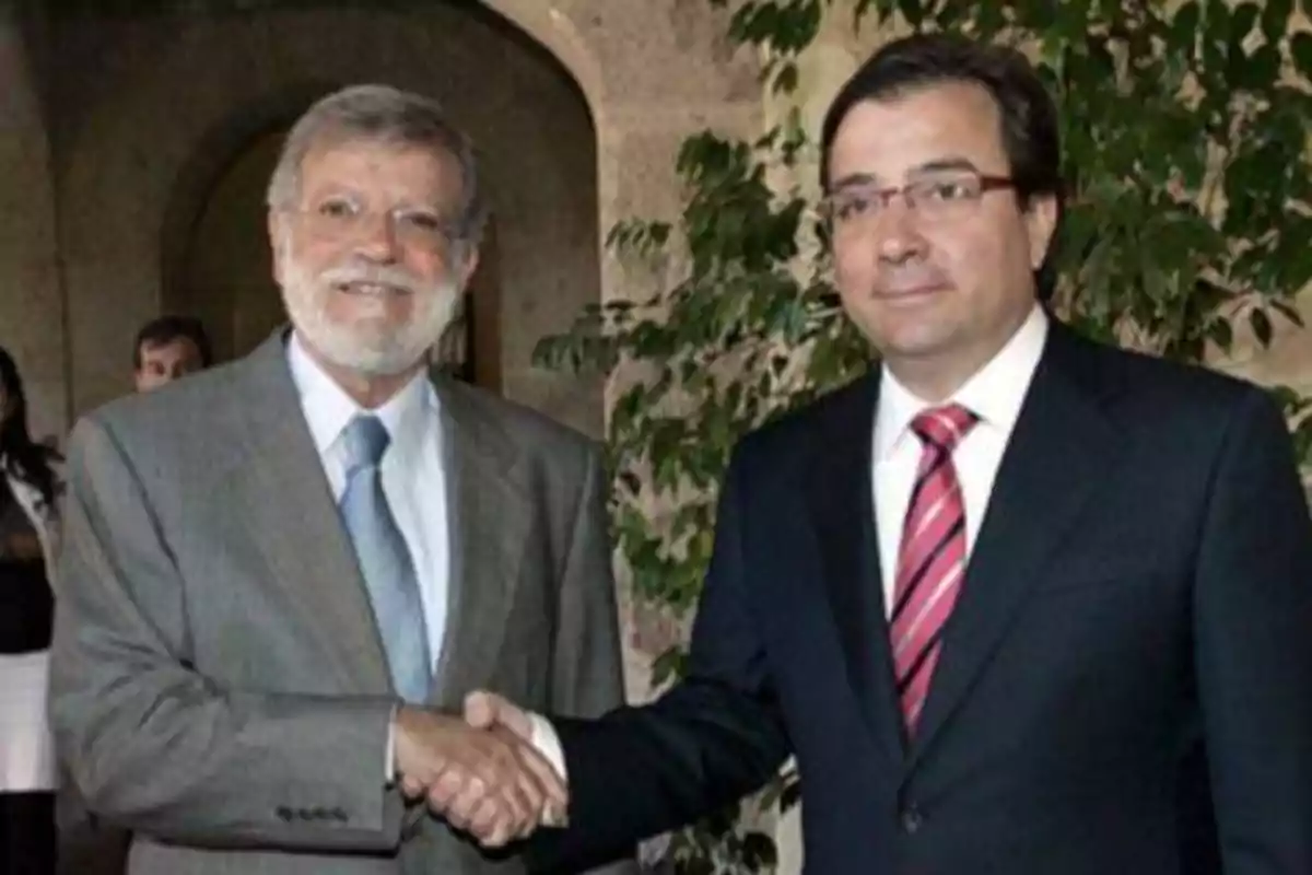 Dos hombres de traje y corbata se dan la mano mientras sonríen, uno de ellos lleva gafas y el otro tiene barba.