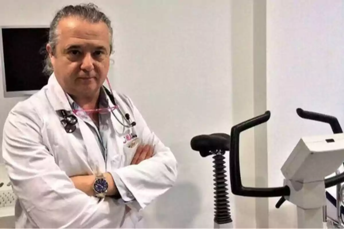 Un médico con bata blanca y estetoscopio cruzado de brazos junto a una bicicleta estática en una consulta médica.