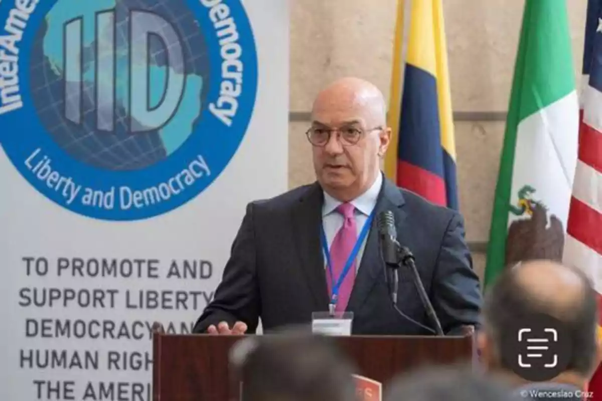 Un hombre de traje y corbata rosa habla en un podio con un micrófono, detrás de él hay un cartel con el logo de "InterAmerican Institute for Democracy" y varias banderas.