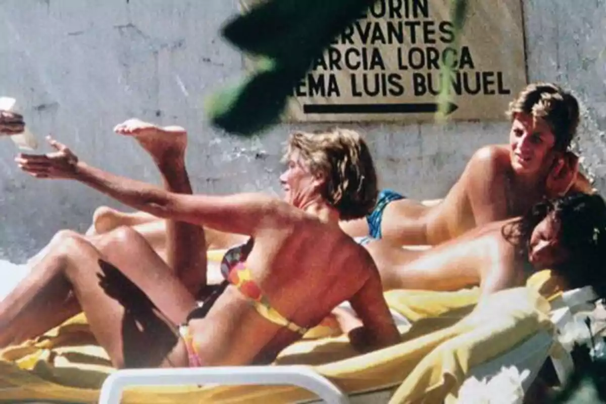Personas en trajes de baño descansando en tumbonas al aire libre con un cartel en la pared de fondo.