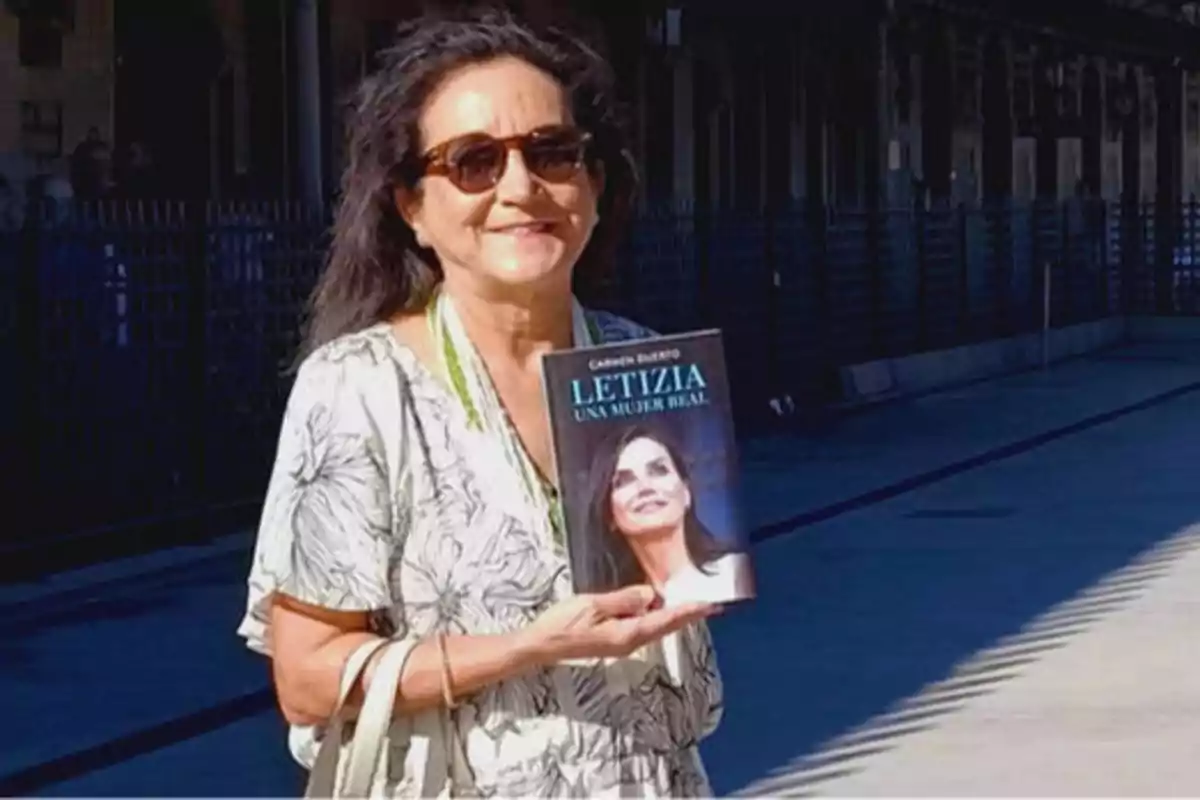 Una mujer con gafas de sol sostiene un libro titulado "Letizia: Una mujer real" mientras sonríe a la cámara.