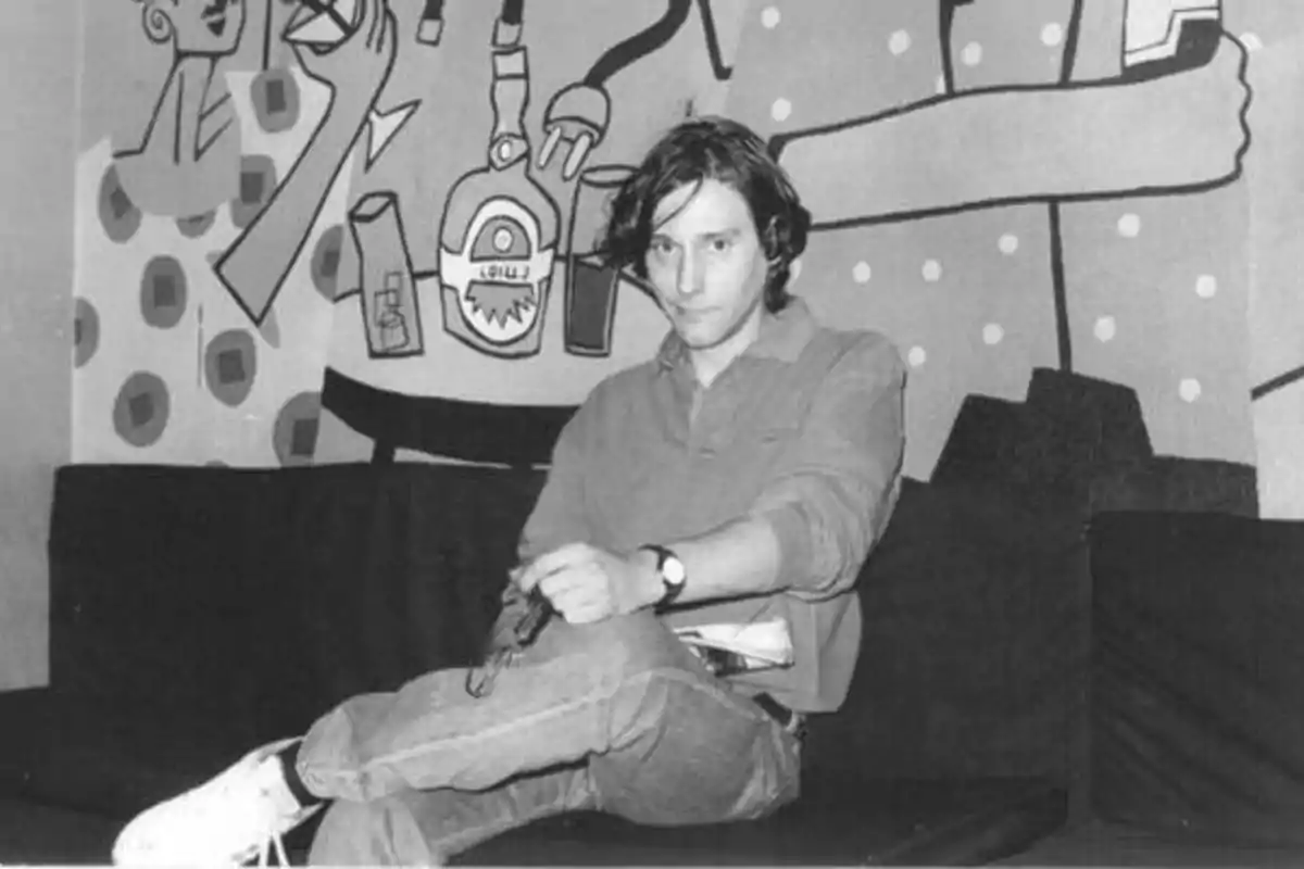 Un hombre joven sentado en un sofá con las piernas cruzadas, sosteniendo un objeto en sus manos, con un mural artístico en el fondo.