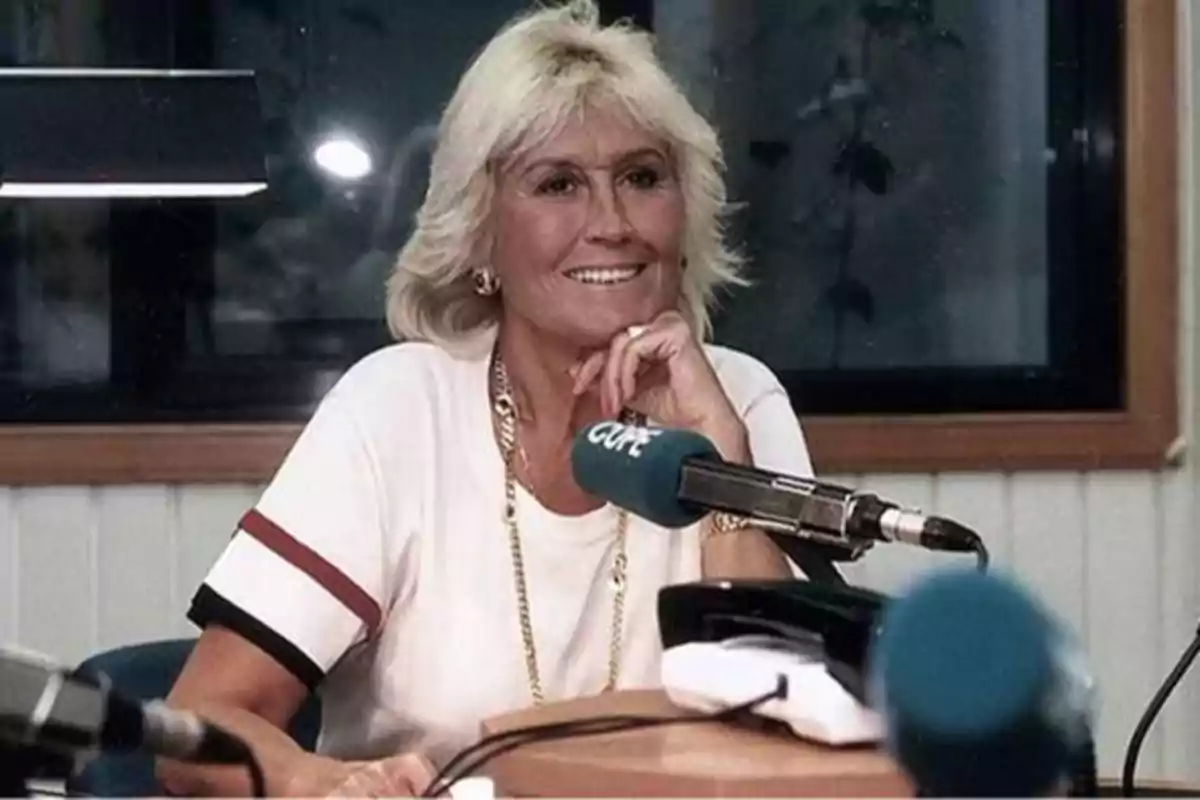 Una mujer de cabello rubio y corto está sentada frente a un micrófono en un estudio de radio, sonriendo y apoyando su barbilla en su mano.