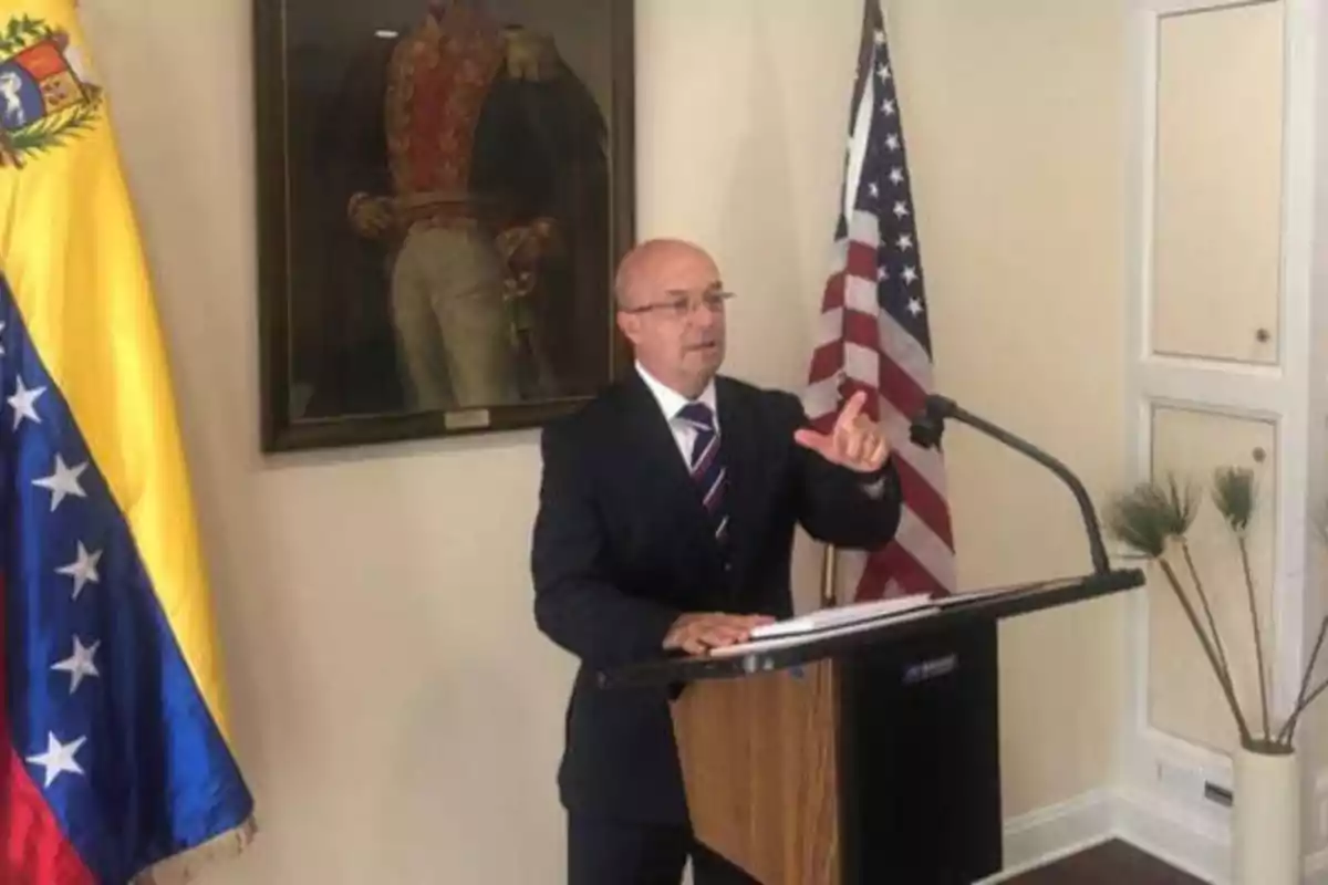 Un hombre de traje y corbata habla en un podio con las banderas de Venezuela y Estados Unidos detrás de él y un retrato de un militar en la pared.