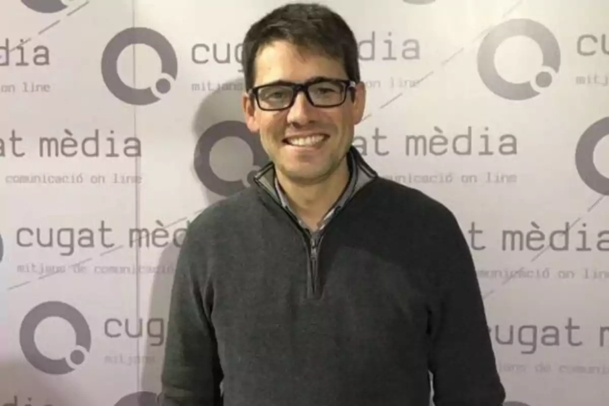 Un hombre con gafas y suéter gris sonríe frente a un fondo con el logotipo y el nombre de "Cugat Mèdia".