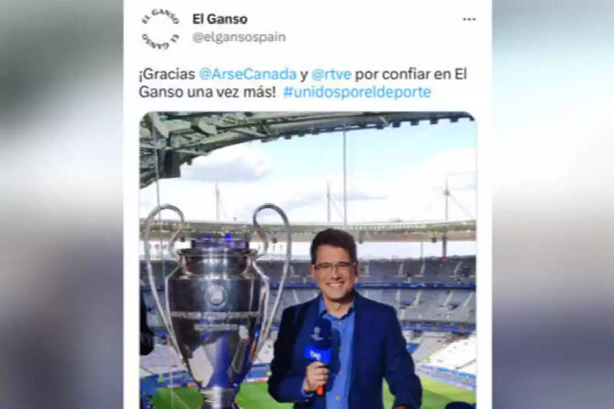 Un hombre con gafas y un micrófono de RTVE está de pie junto a un trofeo grande en un estadio de fútbol vacío.