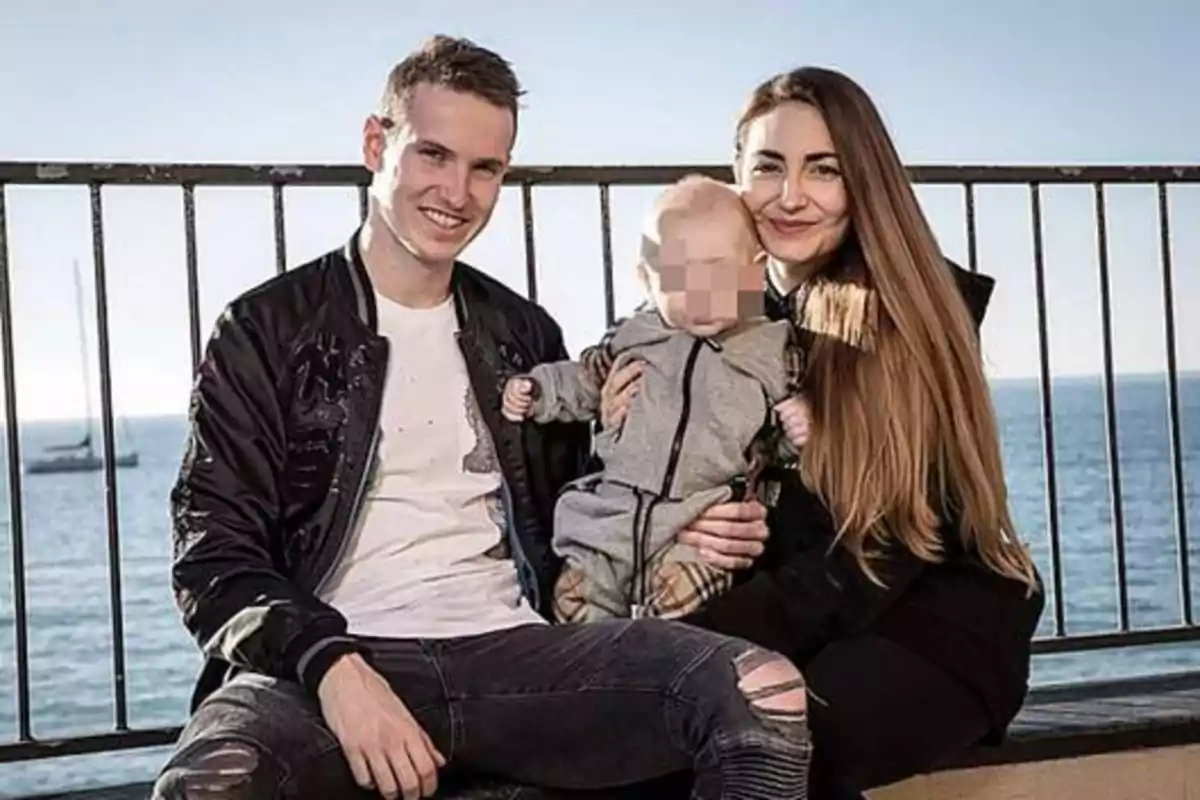 Una pareja sonriente sentada en un banco al aire libre sosteniendo a un bebé con el mar de fondo.