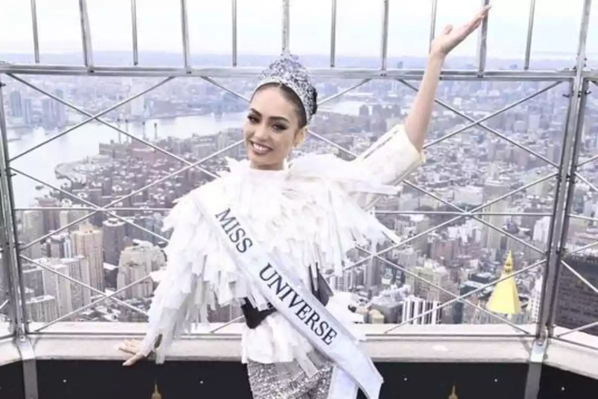 Una mujer con una corona y una banda que dice "Miss Universe" posa sonriente frente a una vista panorámica de una ciudad.