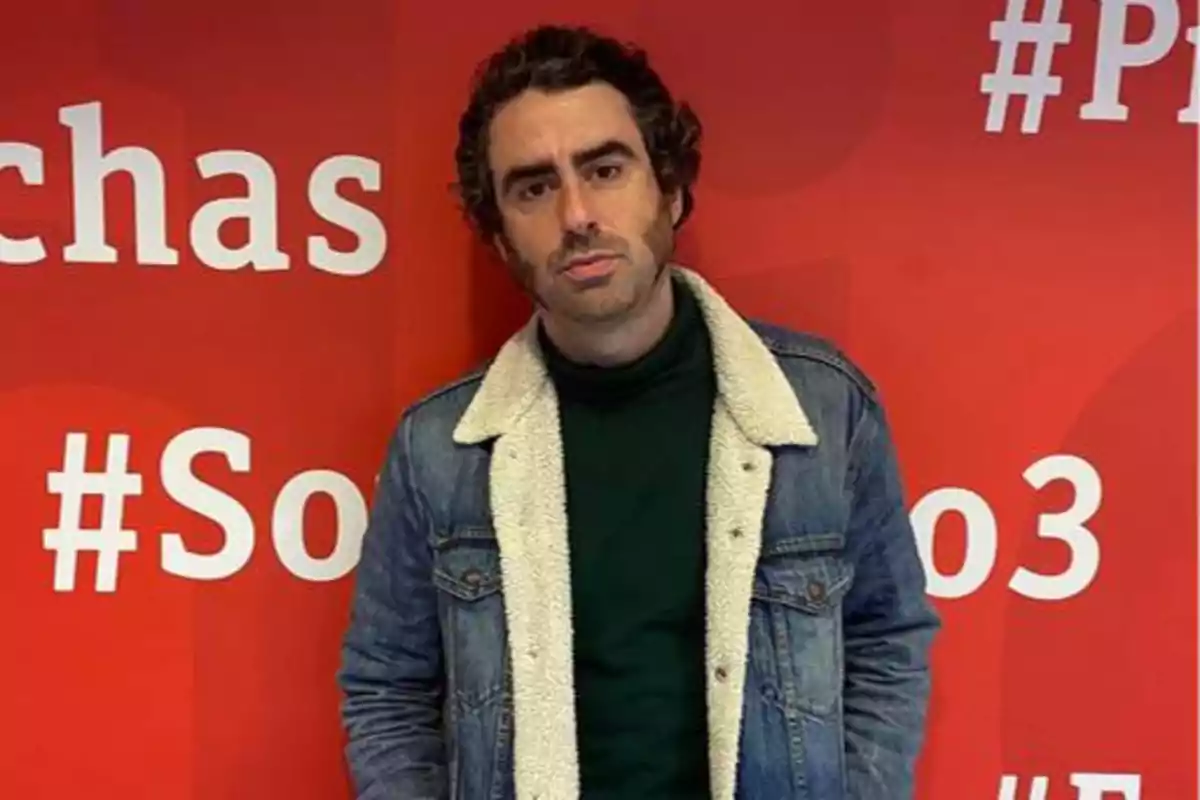 Un hombre con chaqueta de mezclilla y suéter verde oscuro posando frente a una pared roja con hashtags en letras blancas.