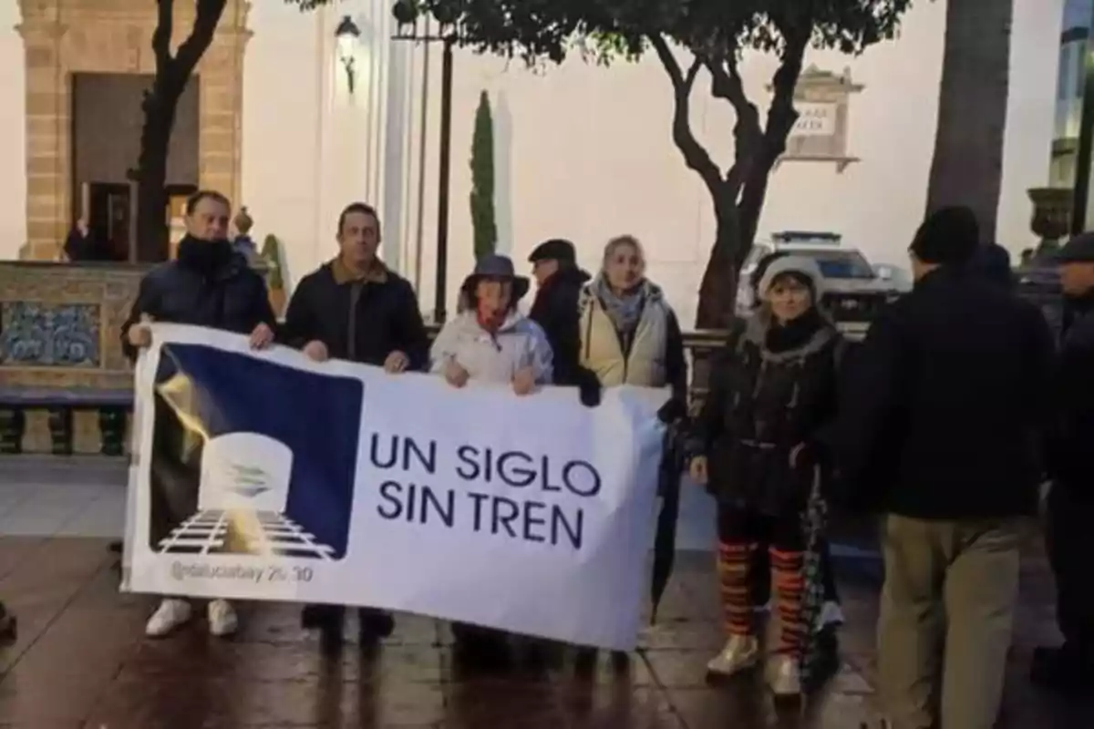 Un grupo de personas sostiene una pancarta que dice "UN SIGLO SIN TREN" en una plaza pública.