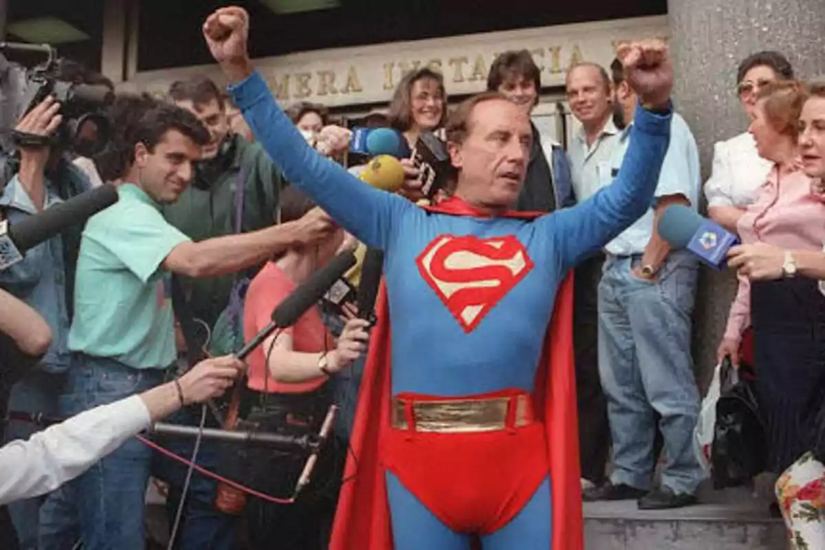 Un hombre disfrazado de superhéroe con una "S" en el pecho levanta los brazos mientras es rodeado por periodistas y cámaras.